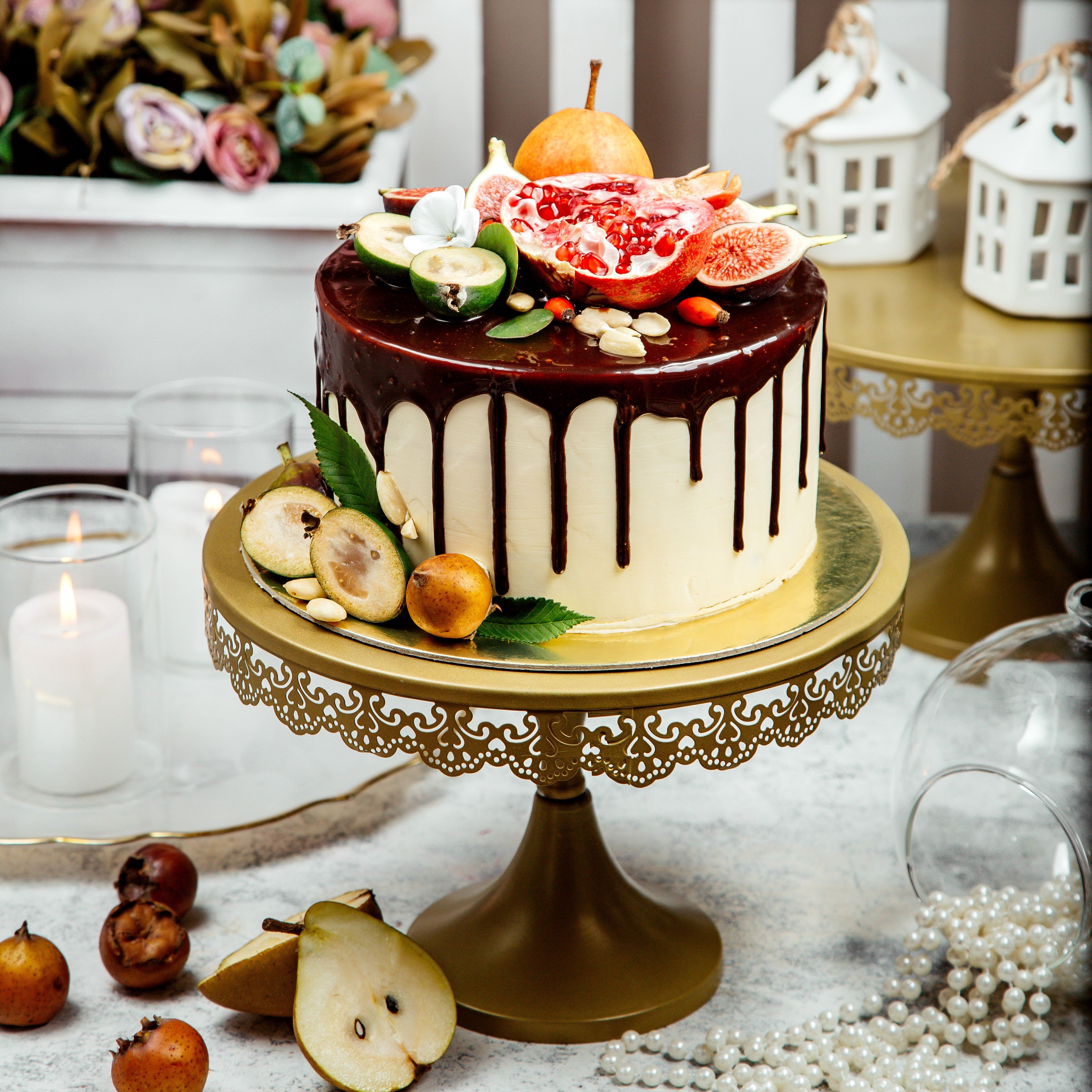 Decorative cake with chocolate drizzle on a gold stand, surrounded by fruits and flowers.
