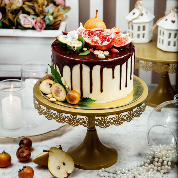Decorative cake with chocolate drizzle on a gold stand, surrounded by fruits and flowers.