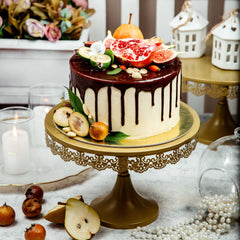 Decorative cake with chocolate drizzle on a gold stand, surrounded by fruits and flowers.